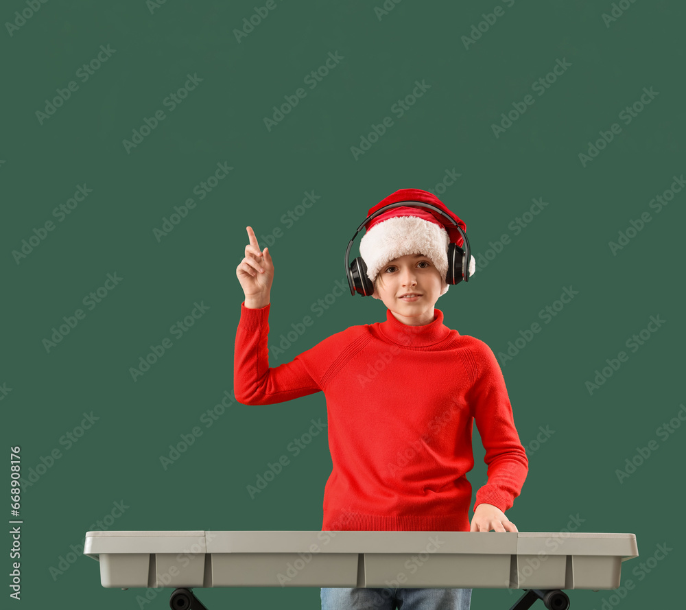 Cute little boy in Santa hat with headphones and synthesizer pointing at something on green background