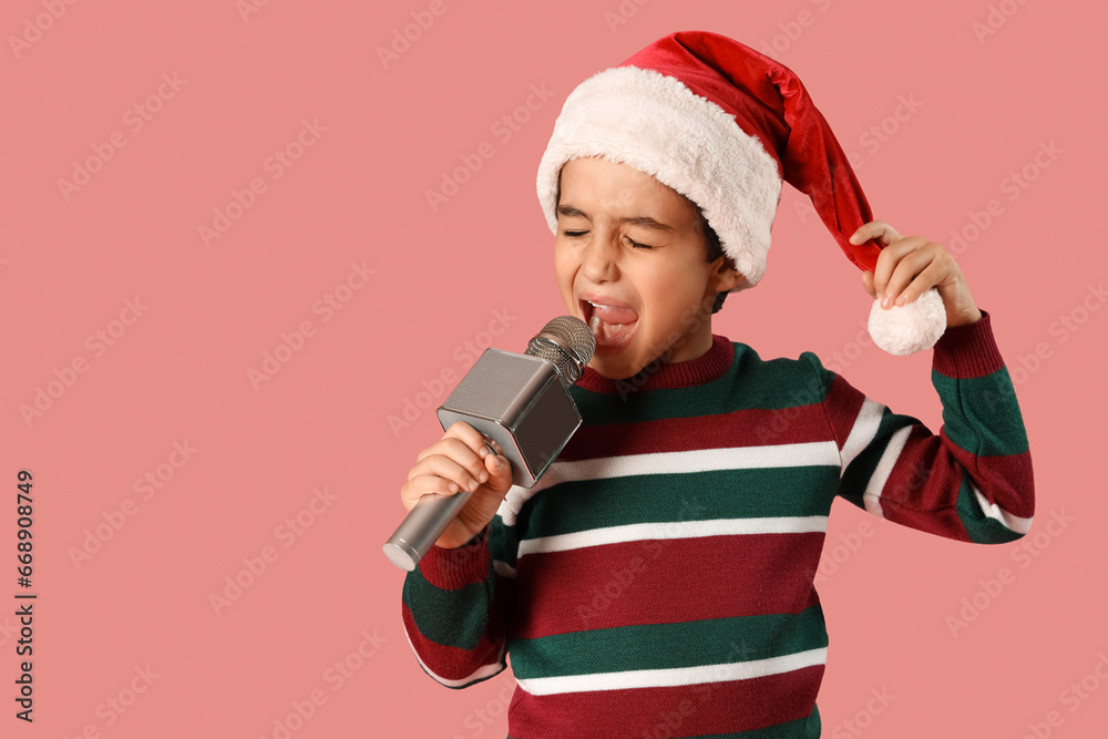 Cute little boy in Santa hat with microphone singing on pink background