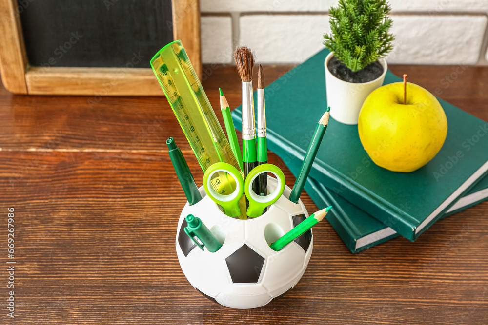 Holder in shape of soccer ball with different stationery, tasty apple and chalkboard on wooden desk, closeup