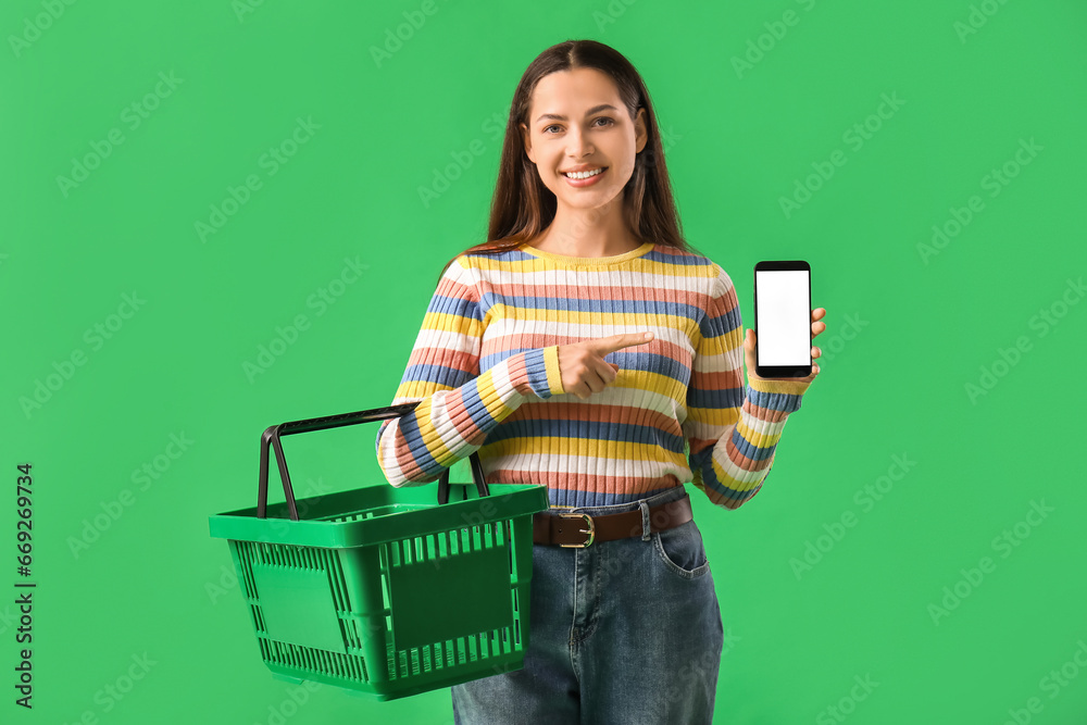 Young woman with shopping basket pointing at mobile phone on green background