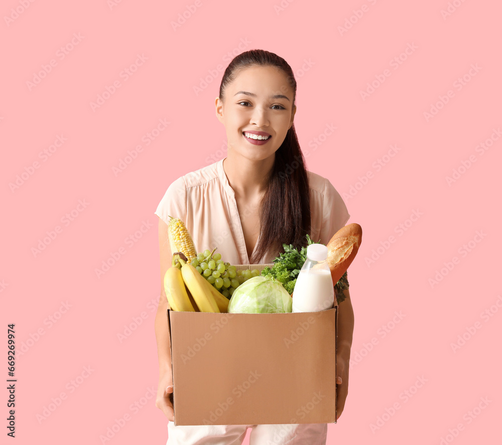 Young Asian woman with shopping box full of fresh food on pink background