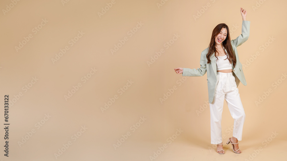 Young Asian happy smiling woman in casual business suit raising her hands up like a winner isolated over beige background - with empty copy space - in full body