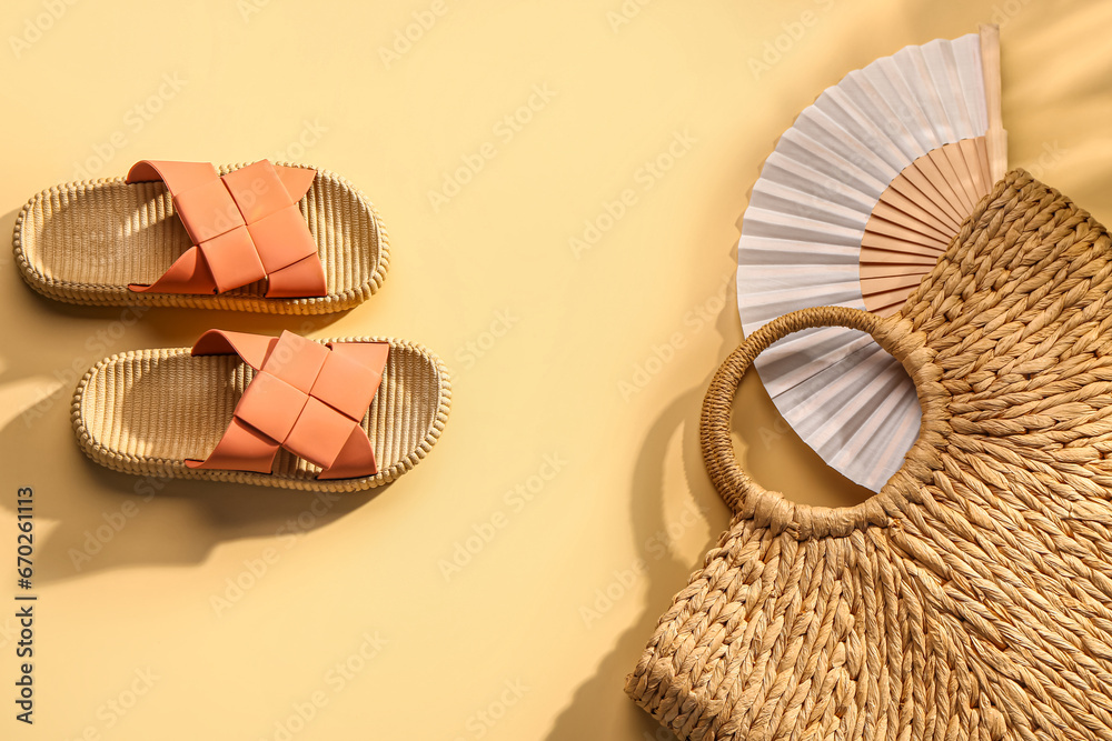 Female stylish flip flops with wicker bag and fan on pale yellow background