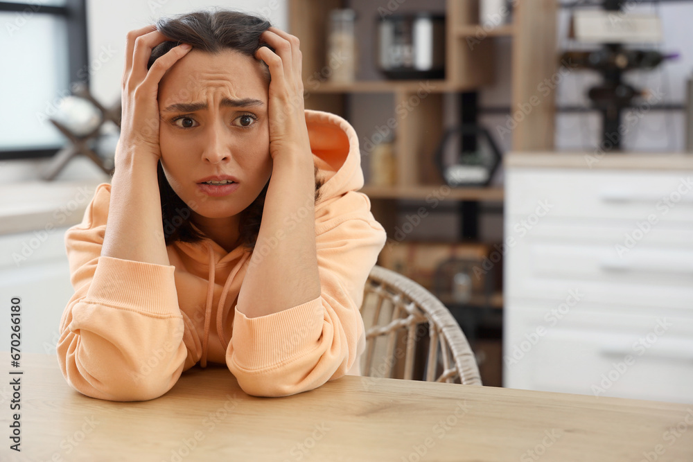 Beautiful young afraid woman sitting at table in kitchen