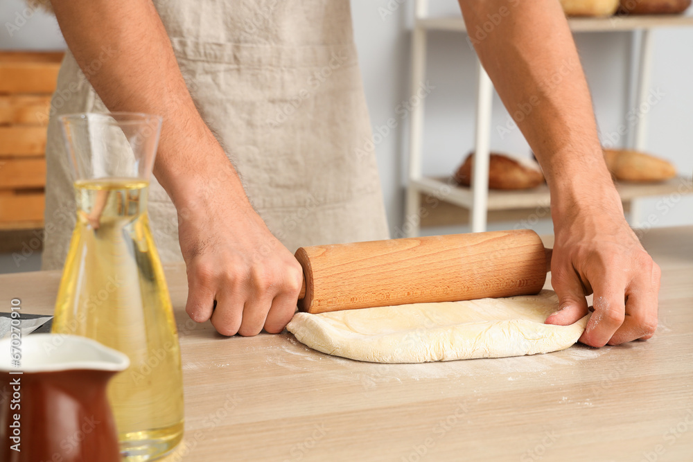 Young handsome man rolling out dough on table in bakery