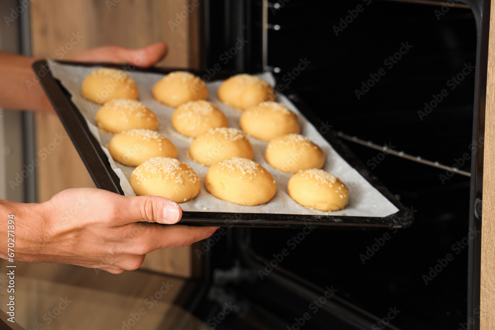 Handsome young chef putting baking tray with tasty buns into oven, closeup