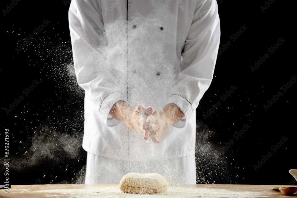 Young chef clapping and sprinkling flour over dough on table against black background