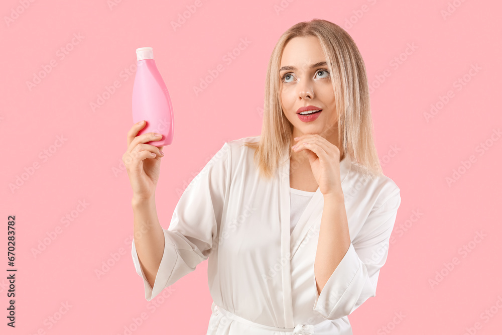 Thoughtful young blonde woman with bottle of hair product on pink background