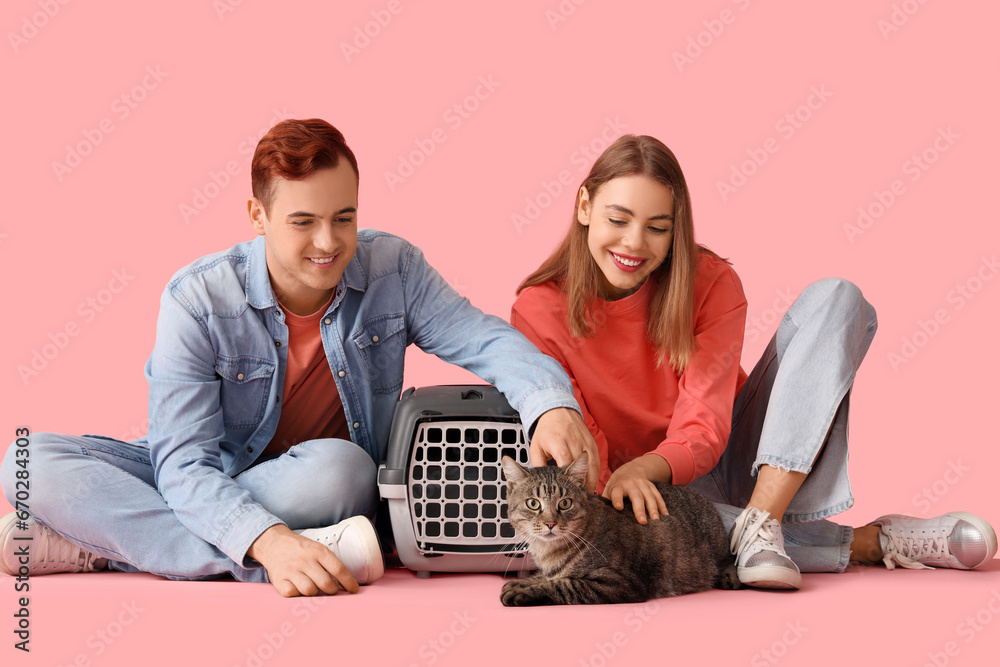 Young couple with cute cat and carrier on pink background