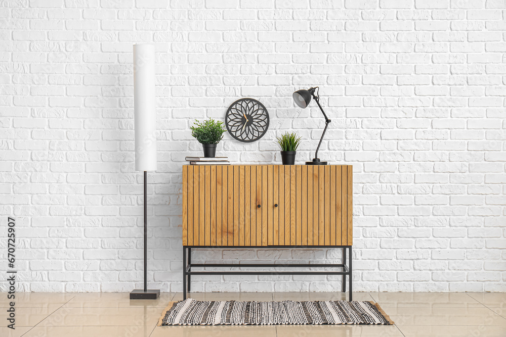 Wooden cabinet with desk lamp and houseplants near white brick wall