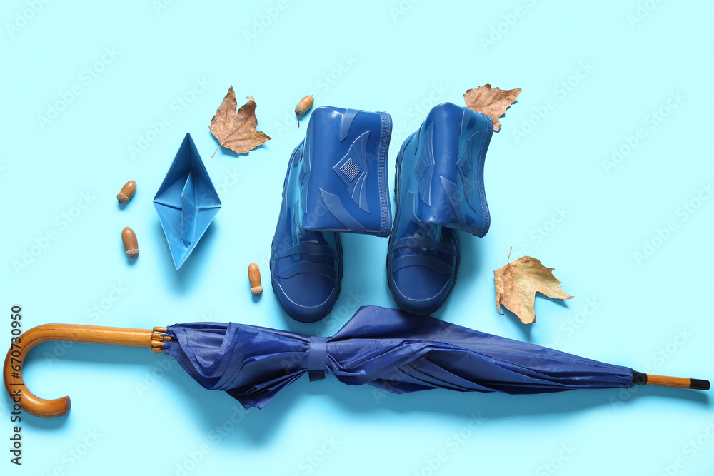 Blue gumboots with autumn leaves, paper ship and umbrella on color background