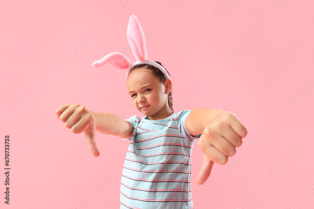 Little girl in bunny ears showing dislike on pink background