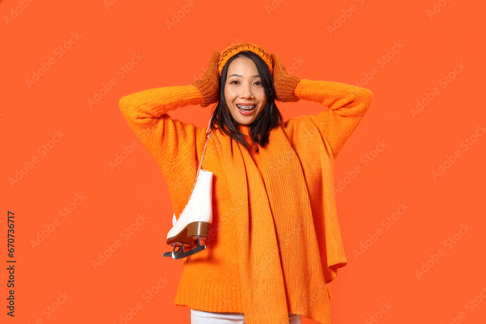 Young Asian woman in winter clothes with ice skates on orange background