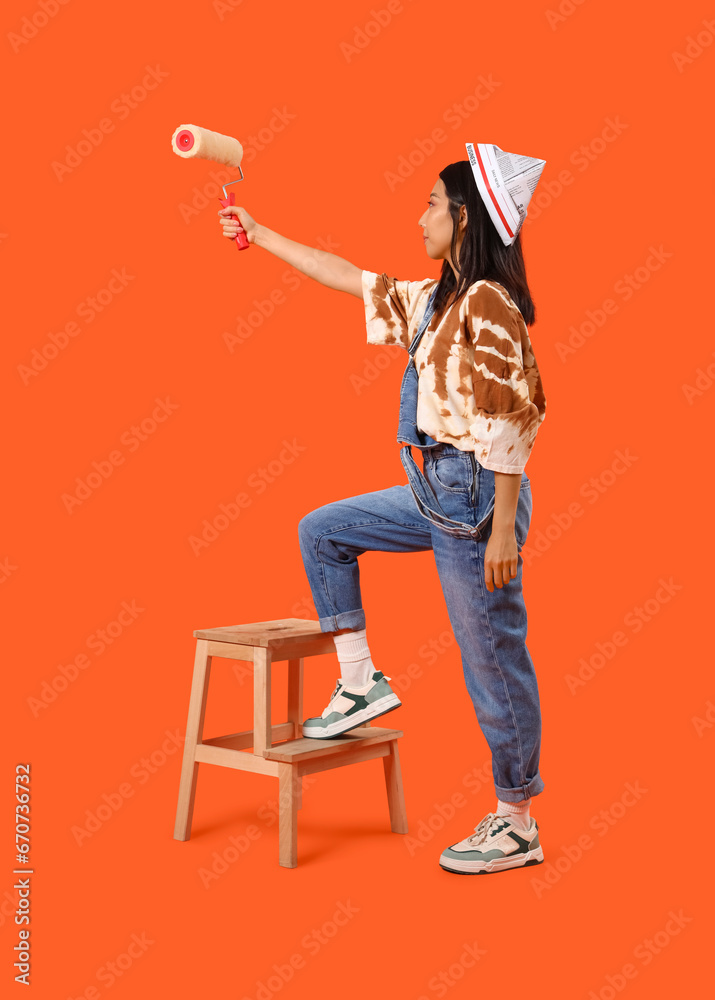 Young Asian woman with paint roller on orange background