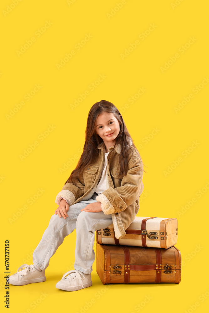 Little girl in winter clothes sitting on suitcases against yellow background