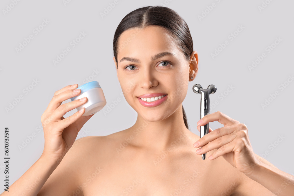 Beautiful young woman with facial massage tool and cream on light background