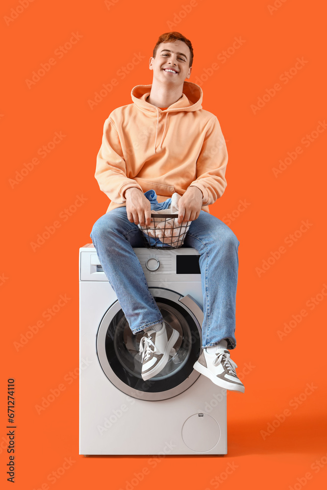 Young man with laundry sitting on washing machine against orange background