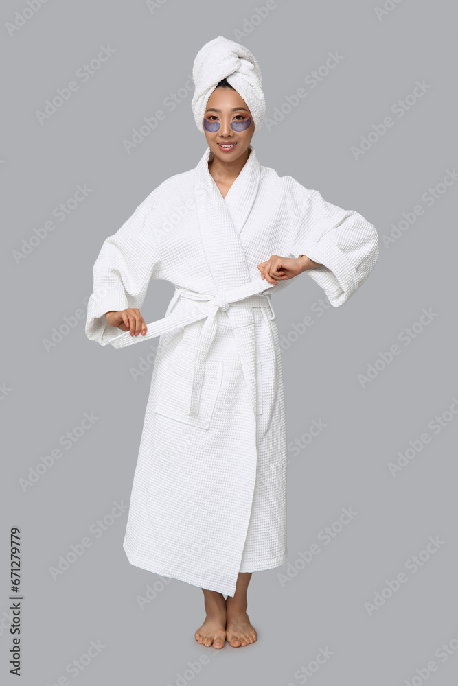 Young Asian woman in bathrobe with under-eye patches on light background