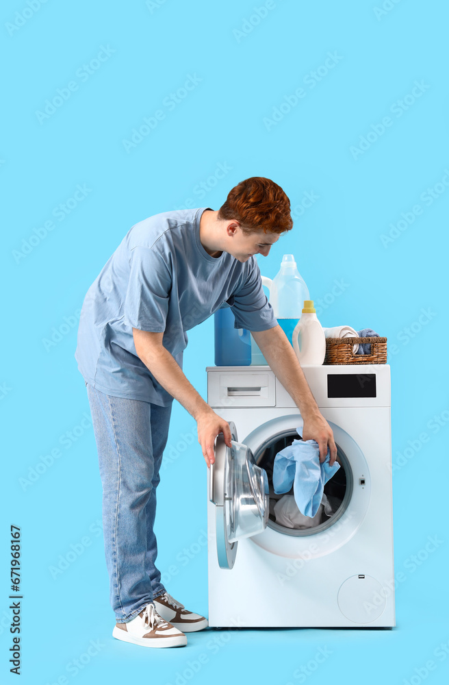 Young man putting laundry into washing machine on blue background