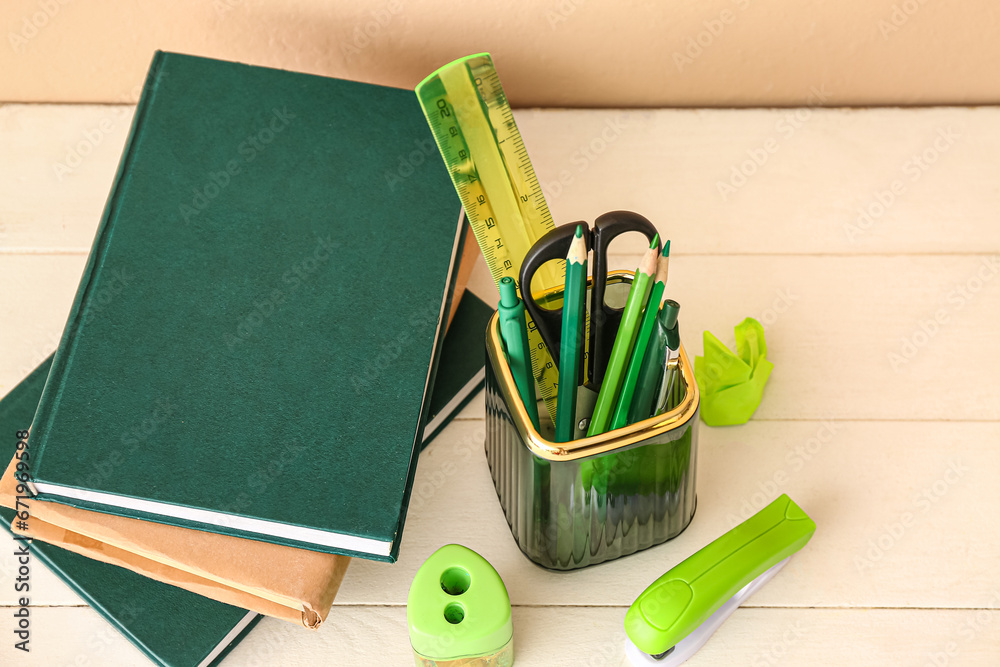 Holder with different stationery and books on light wooden desk, closeup