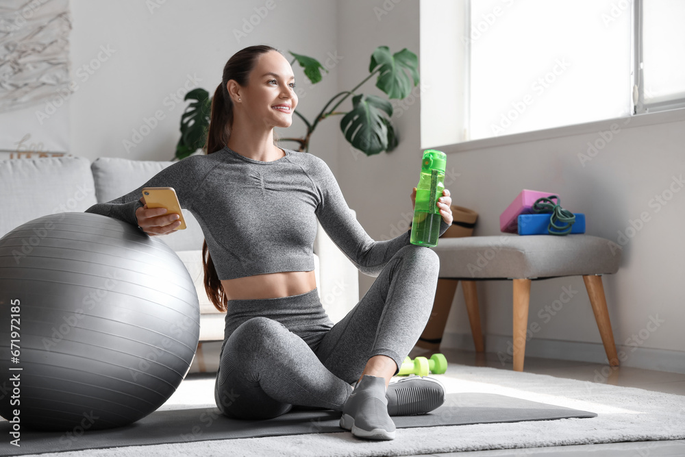 Sporty young woman with fitball, mobile phone and water bottle at home