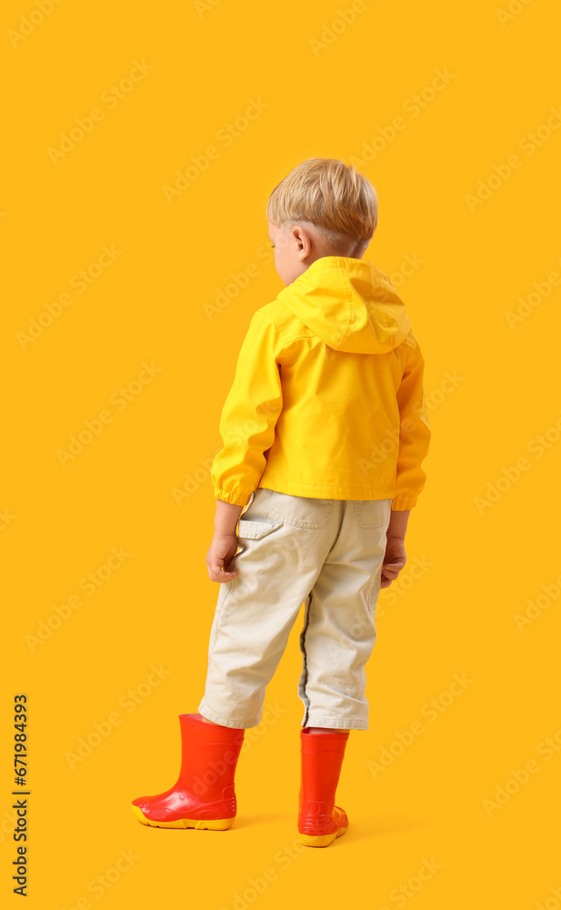 Little boy in rubber boots on yellow background, back view