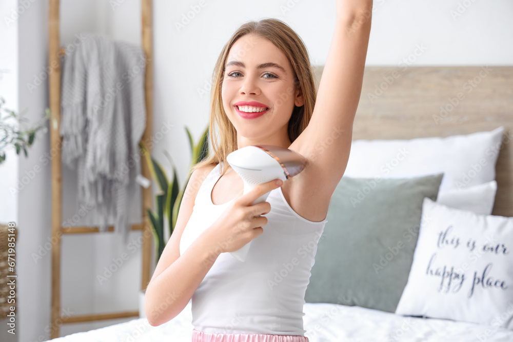 Young woman epilating her armpits with modern photoepilator in bedroom at home