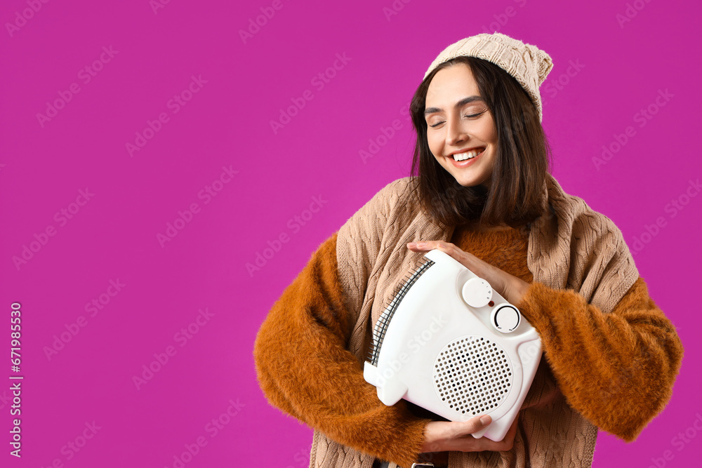 Young woman in winter clothes with electric heater on purple background