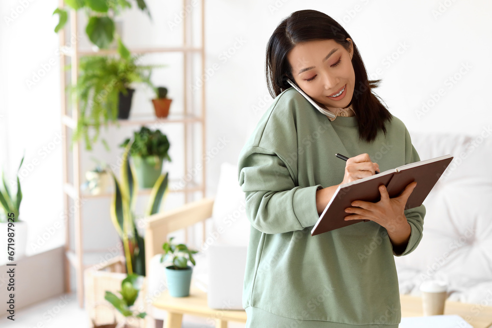 Young Asian woman with notebook talking by mobile phone at home