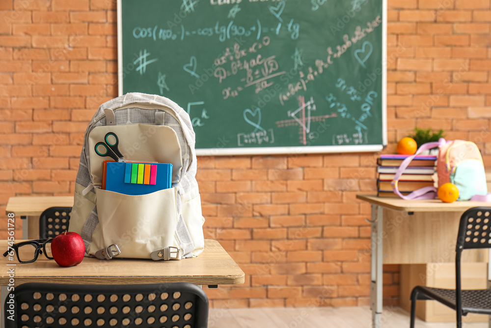 Stylish school backpack with stationery, eyeglasses and apple on desk in classroom