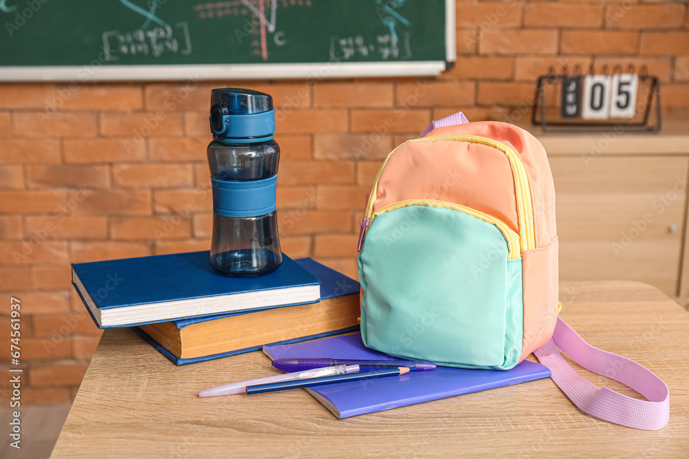 Stylish school backpack with bottle of water and stationery on desk in classroom