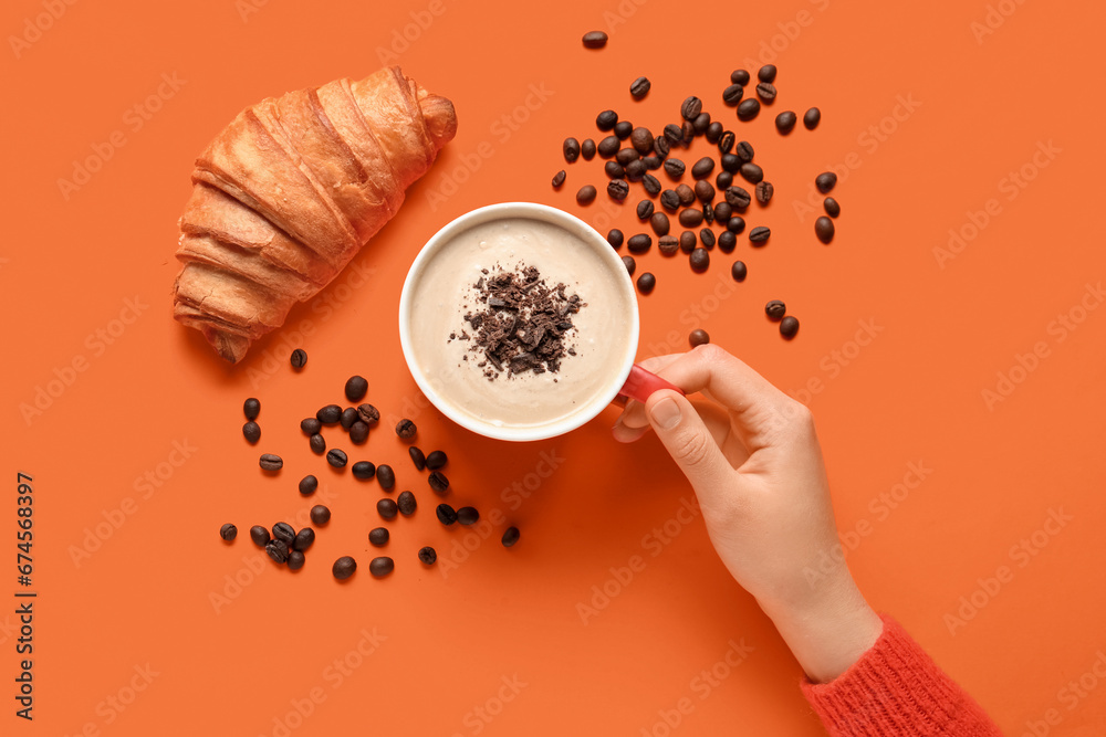 Woman holding cup of hot coffee with chocolate and croissant on orange background