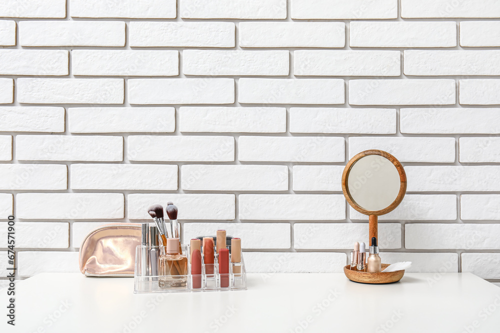 Holder with different makeup products and mirror on dressing table near light brick wall in room