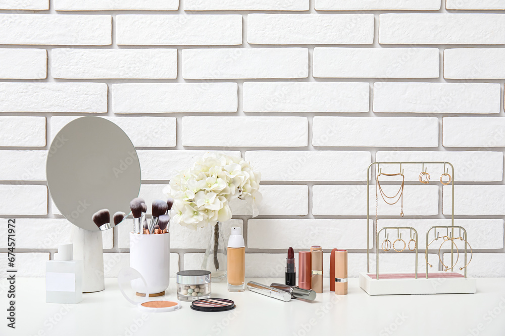 Different makeup products, mirror and beautiful flowers on dressing table near light brick wall in room