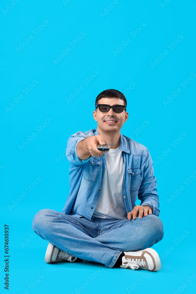 Handsome young man in 3D glasses watching TV while sitting on floor against blue background