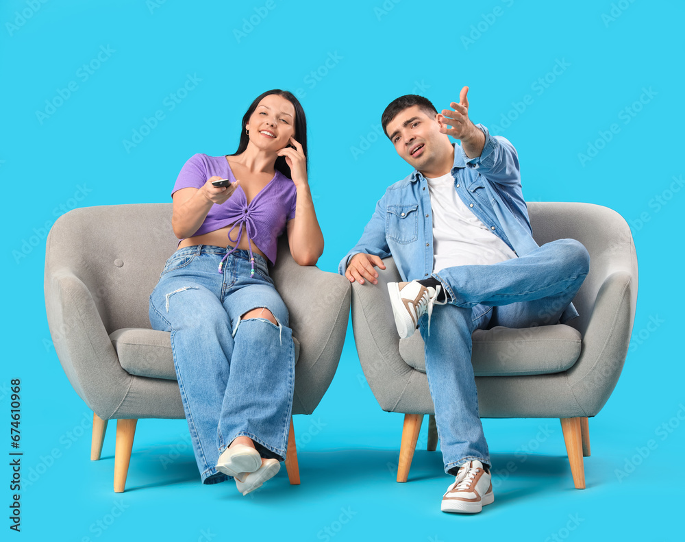 Young couple sitting on armchairs and watching TV together against blue background