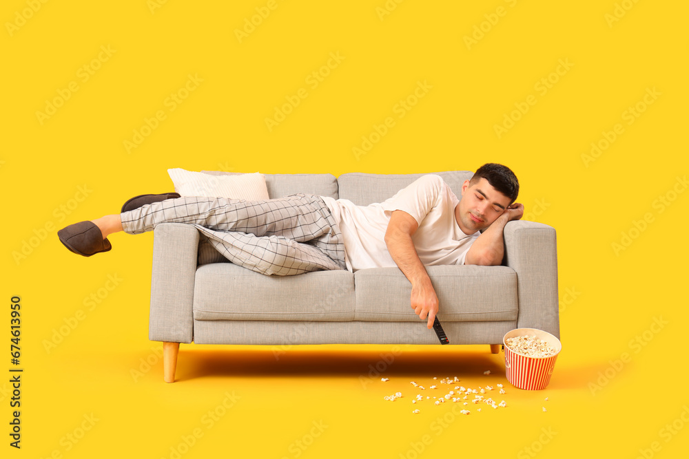 Bored young man sleeping on sofa while holding TV remote control against yellow background