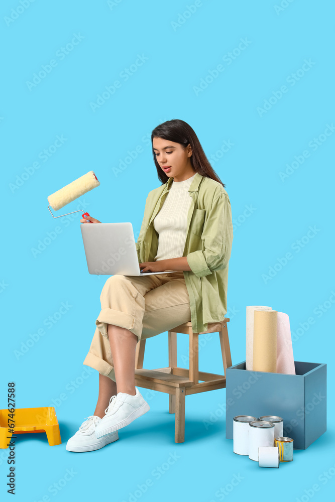 Young woman with paint roller and laptop on blue background