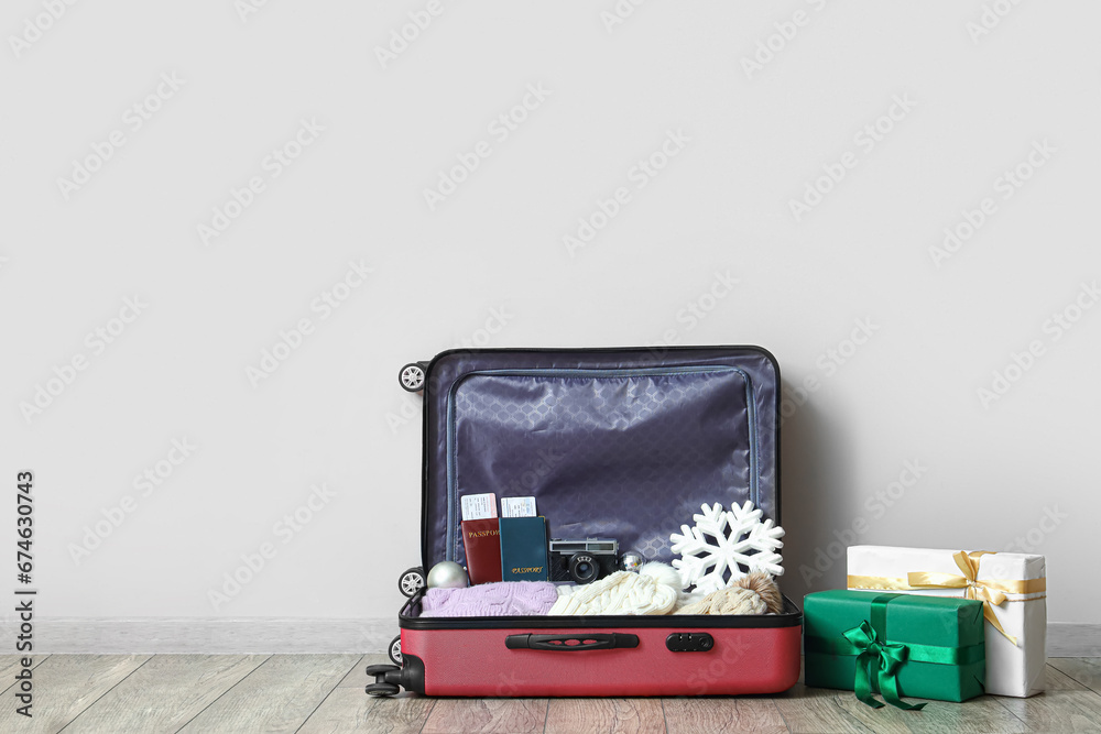 Open suitcase with winter clothes and Christmas gifts near light wall