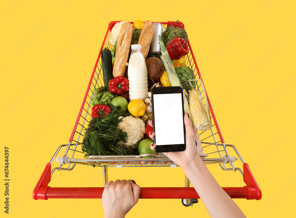 Woman with mobile phone pushing shopping cart full of food on yellow background