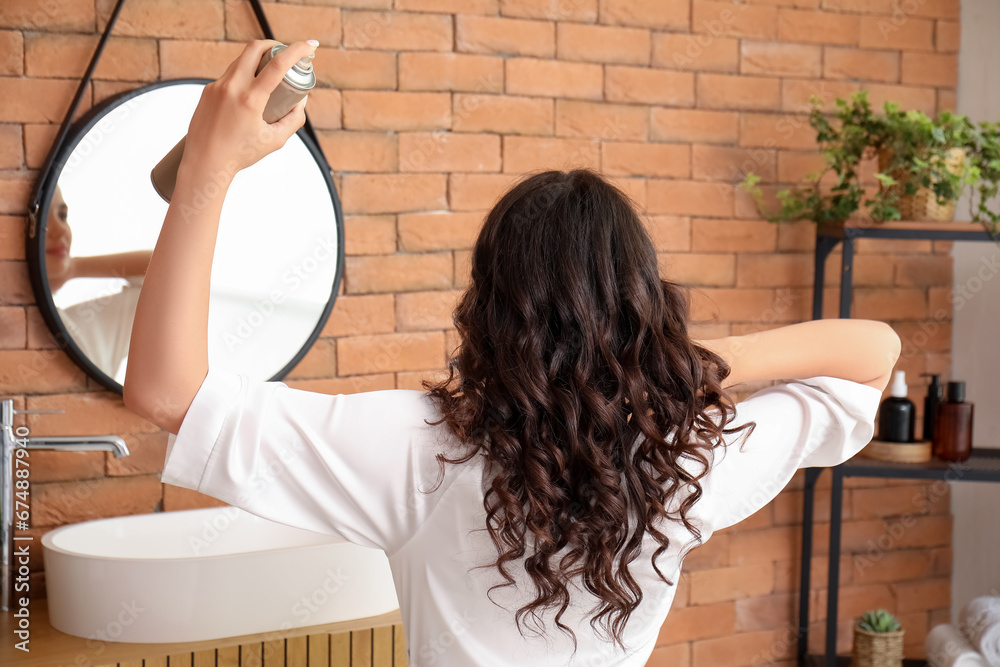 Pretty young woman applying hair spray on her curly hair in bathroom