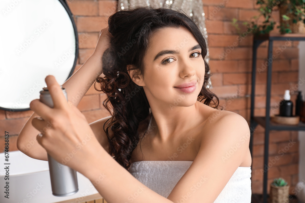 Pretty young woman applying hair spray on her curly hair in bathroom