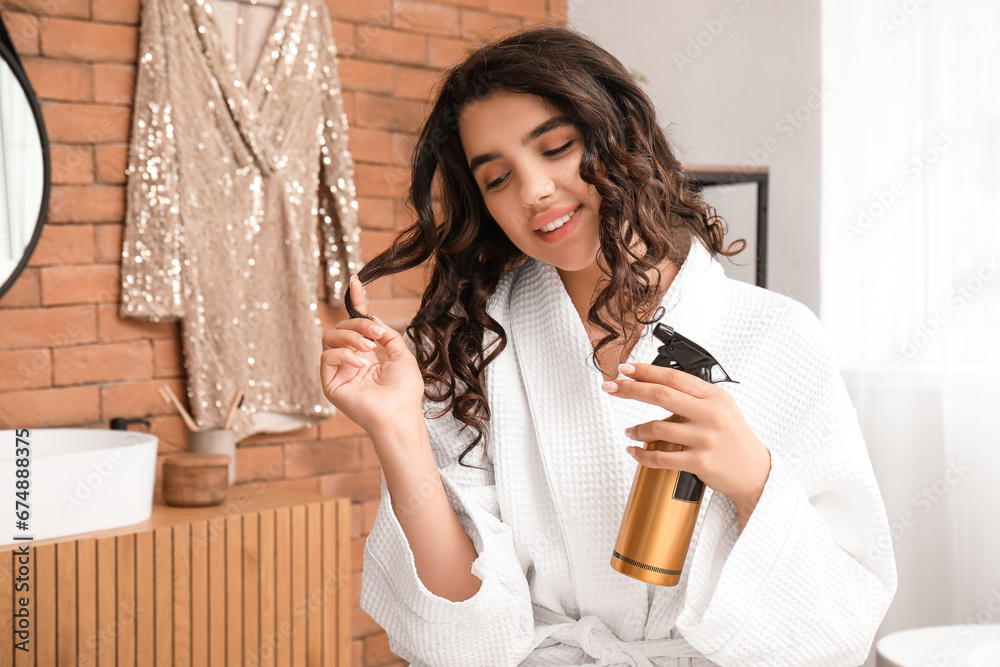 Pretty young woman applying hair spray on her curly hair in bathroom