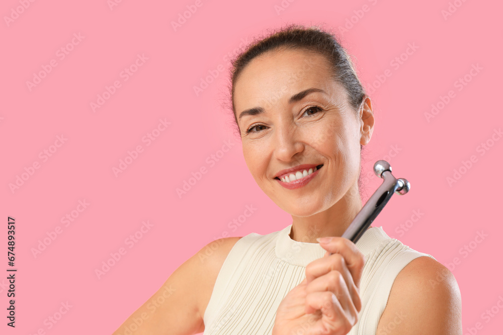 Beautiful mature woman using face massage roller on pink background