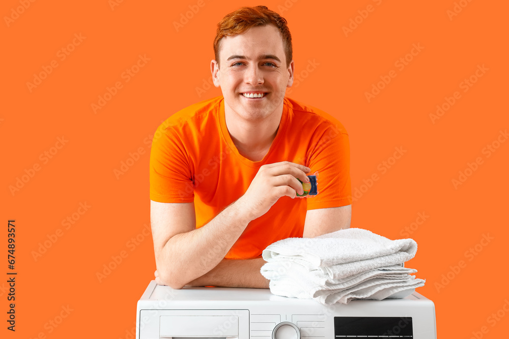 Young man with laundry capsule and towels on orange background