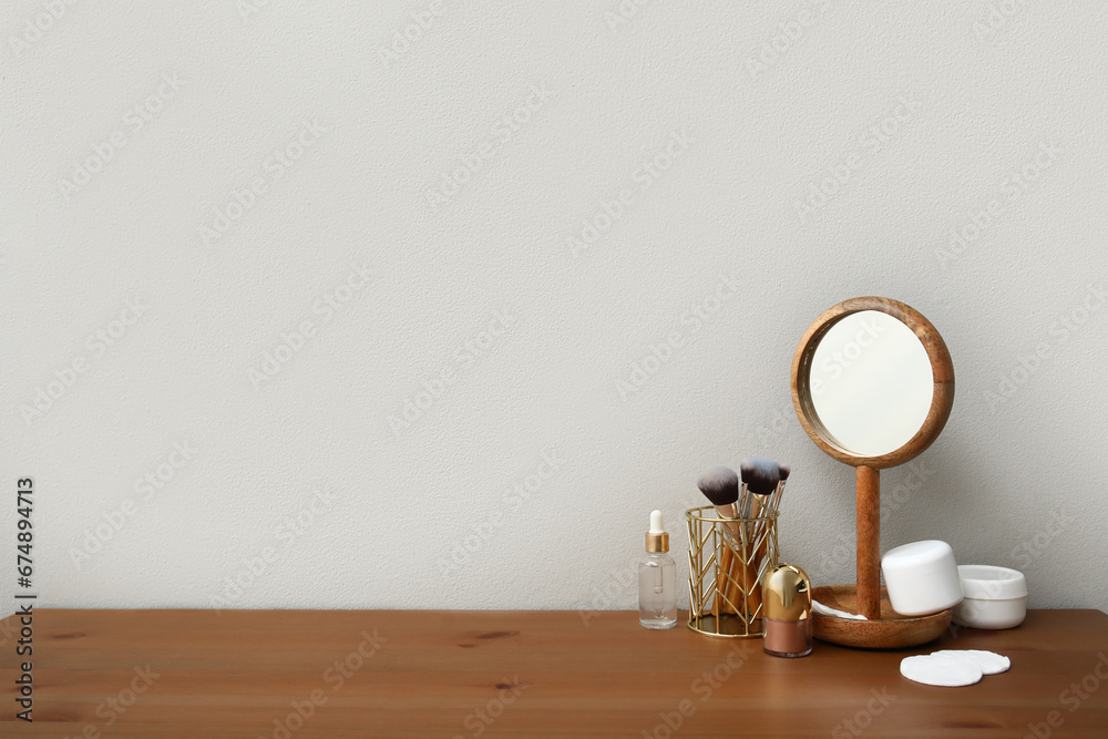 Different makeup products and mirror on dressing table near light wall in room