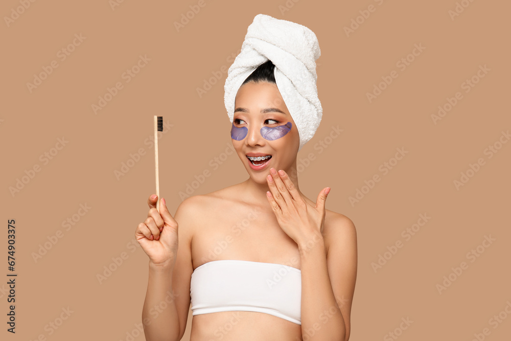 Surprised young Asian woman with under-eye patches and toothbrush on brown background