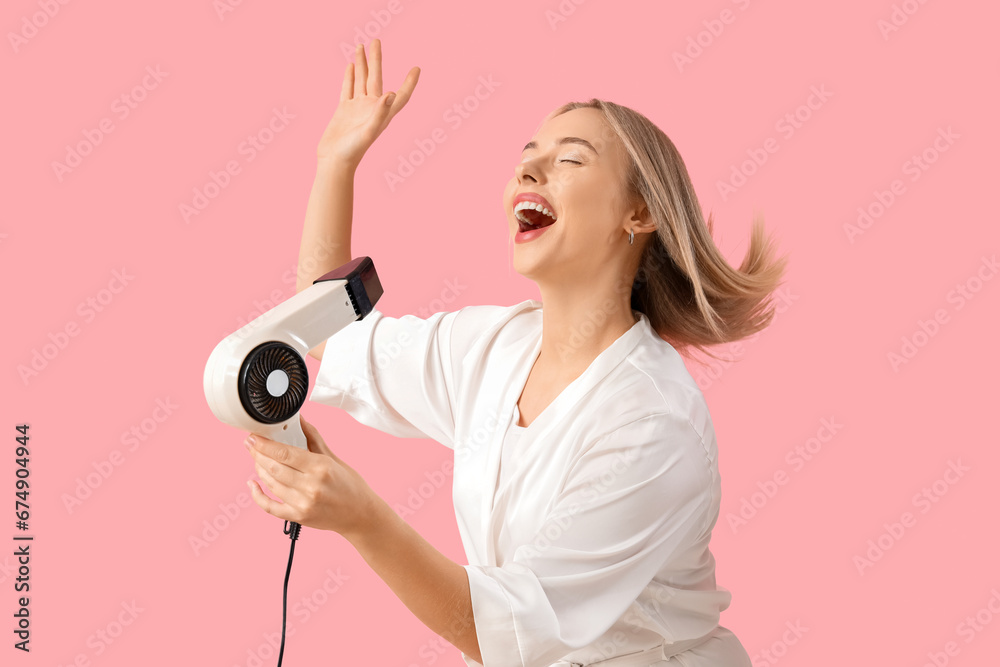 Young blonde woman drying hair on pink background