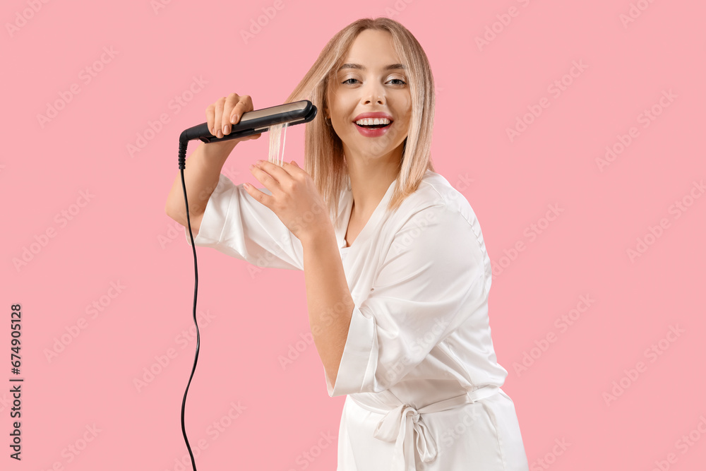 Young blonde woman straightening hair on pink background