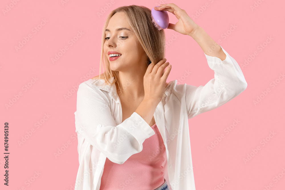 Young blonde woman brushing hair on pink background